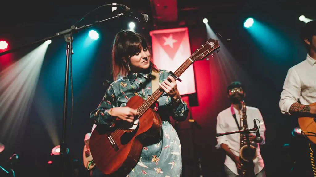 A woman plays an acoustic guitar on stage with a saxophonist and other musicians under colorful lights, while a flag featuring a star hangs in the background—capturing a perfect moment for Music Analytics.
