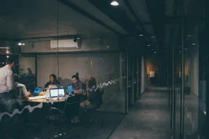 A group of people sit around a conference table with laptops in a glass-walled office, discussing music industry guides, while one person stands nearby; a hallway is visible outside the room.
