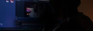 Person editing a video on a computer; the screen displays video editing software with a paused frame of a person wearing glasses and a hoodie, working on an About Symphonic project.