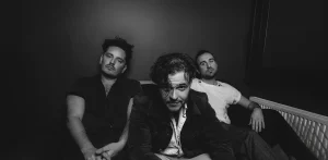 Three men sit closely together on a couch in a dimly lit room, facing the camera with serious expressions. This black and white image evokes the mood of a Protoje Case Study, capturing raw emotion and intensity.