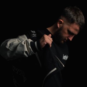 A man with short hair and a beard looks down while adjusting his black and white jacket against a dark background, reminiscent of a scene from a Protoje Case Study.