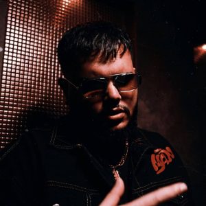 Man wearing sunglasses and a dark jacket with red stitching and a rose design, channeling a cool Protoje Case Study vibe as he stands in a dimly lit room with a textured wall in the background.