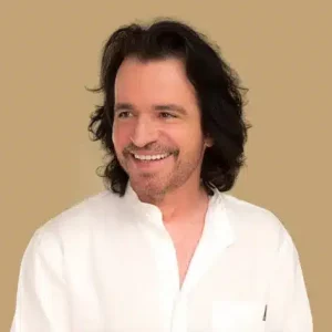 A man with shoulder-length dark hair and a beard, wearing a white shirt, smiles and looks to the side against a plain beige background—capturing a harmonious moment that feels truly About Symphonic.