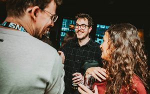Three people are talking and smiling at an indoor Symphonic NEXT social event, drinks in hand. The background is dimly lit with visible windows, creating a lively yet relaxed atmosphere.