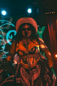 A singer wearing sunglasses, a wide-brimmed hat, and a patterned outfit performs passionately on stage in front of a microphone at Symphonic NEXT.