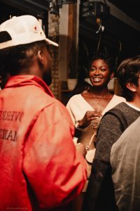 A woman smiles while holding a drink and talking to a man in a red jacket at an indoor Symphonic NEXT social gathering.