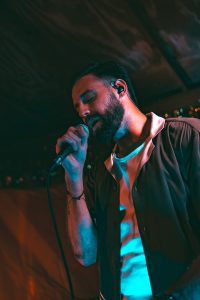 A man with a beard holds a microphone and sings on stage during the Symphonic NEXT event, wearing a white shirt and a dark open shirt, as vibrant stage lighting illuminates him.